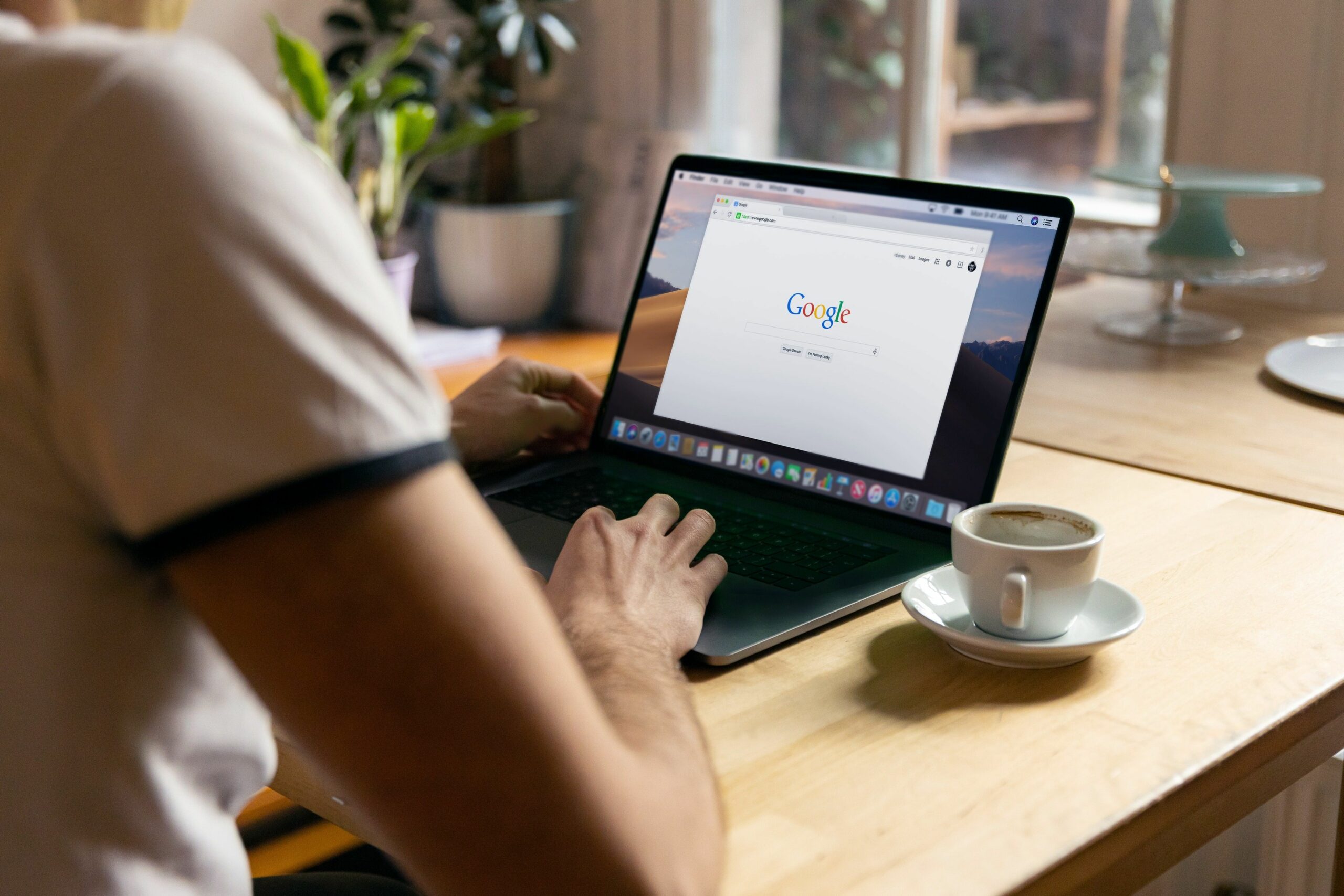 Rückansicht auf einen Menschen am Schreibtisch mit leerer Kaffeetasse und Laptop, auf dem die Startseite von Google zu sehen ist