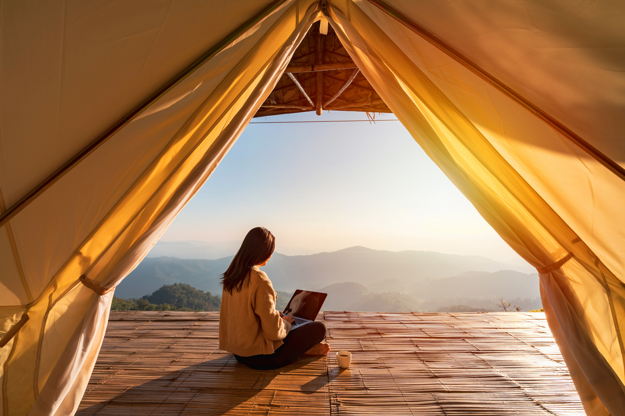 Junge Frau mit Laptop auf dem Schoß sitzt auf einer Plattform mit einer Berglandschaft im Hintergrund und arbeitet